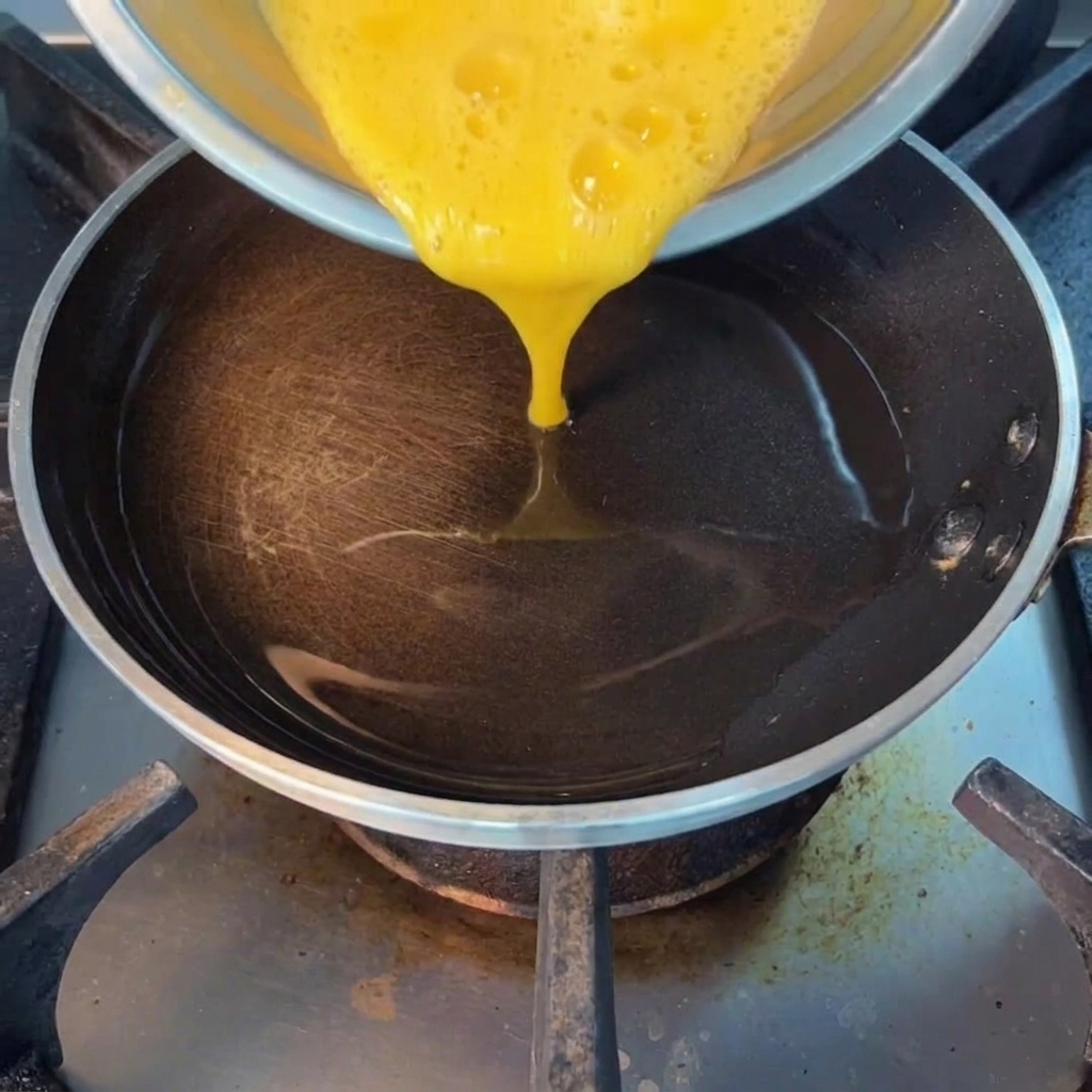 A close-up view of beaten eggs being poured into a hot, oiled pan, ready to be scrambled.