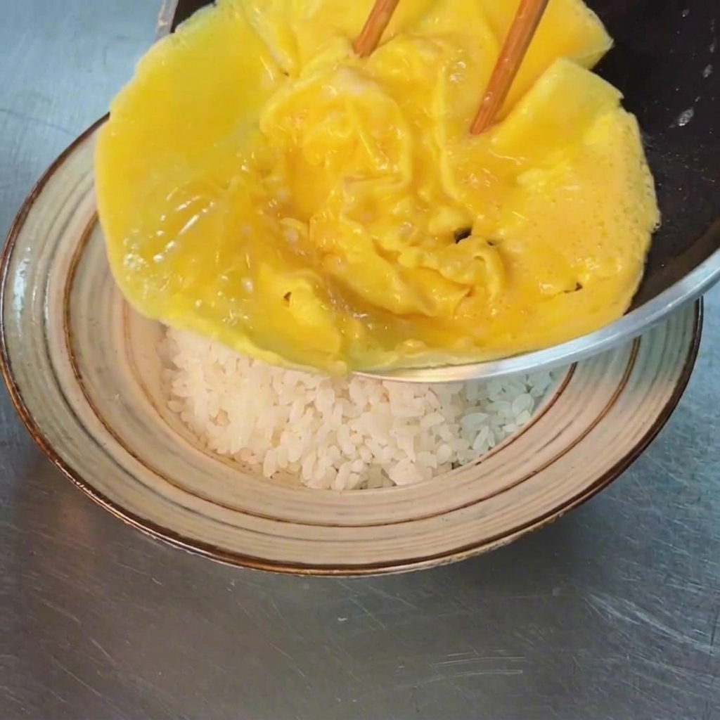 The cooked, soft omelet being carefully slid from a pan onto a mound of white rice in a bowl.