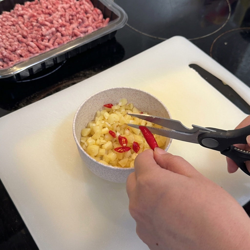 Kitchen shears snipping vibrant red birds eye chilies into a bowl containing chopped garlic.