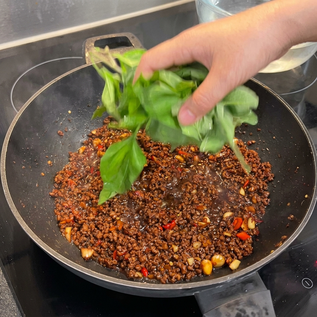 A large handful of fresh holy basil leaves being added to a wok with seasoned beef, ready to be wilted.