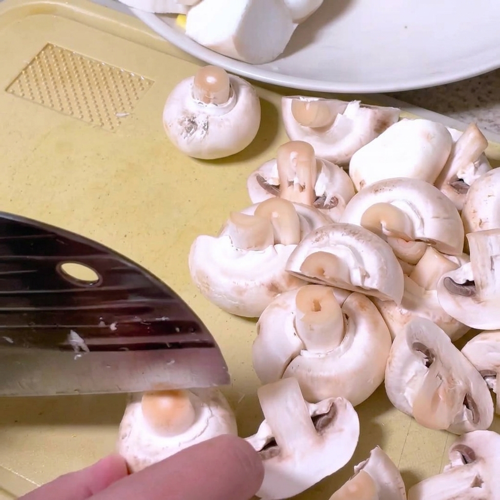 Button mushrooms being halved on a cutting board with a kitchen knife.