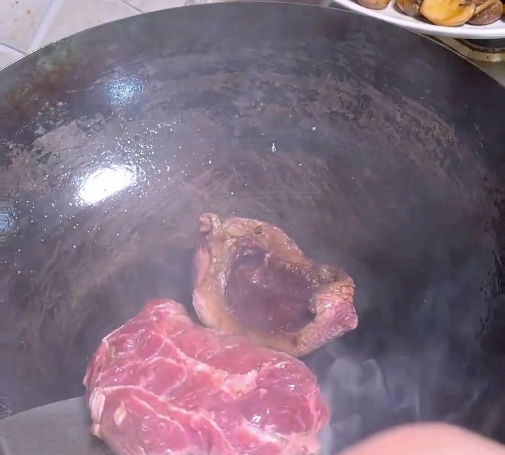 A metal spatula flipping a partially seared steak inside a hot, dark wok.
