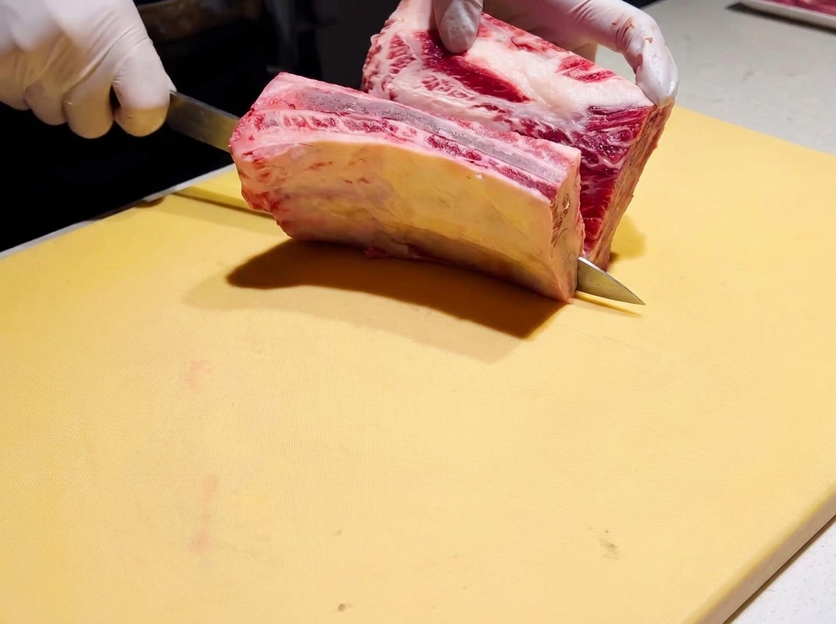 A thick piece of raw beef short rib being butterflied with a chefs knife on a yellow cutting board.