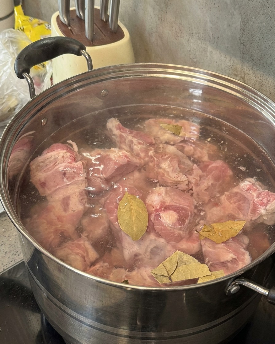 Soju being poured from a green bottle into a pot containing pork bones and bay leaves.