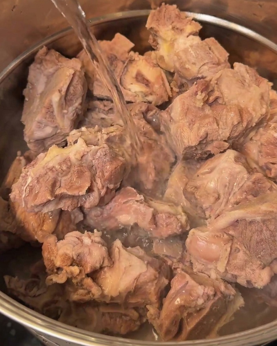 Fresh water being poured into a large metal pot containing the rinsed pork bones.