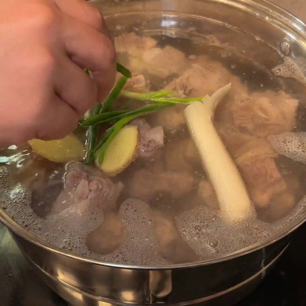 A close-up shot of large white onion chunks and green onion stalks being added into the pot containing parboiled pork bones.