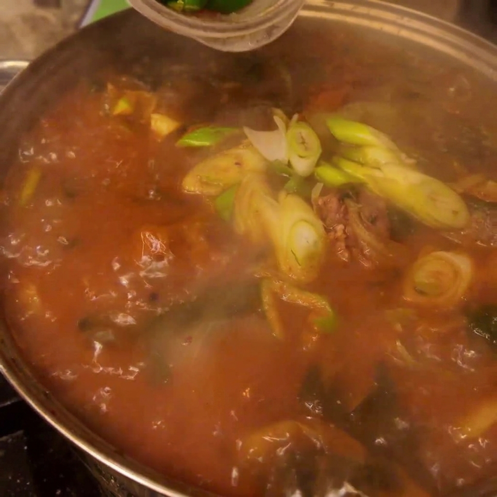 Freshly sliced green and red chili peppers and green onions being sprinkled into a large pot of boiling Gamjatang.