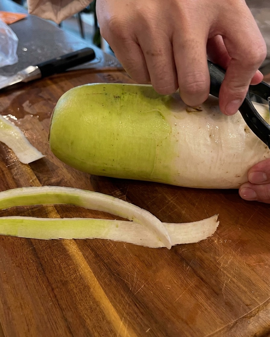 Hands using a black vegetable peeler to peel the green and white skin off a large radish on a wooden cutting board.