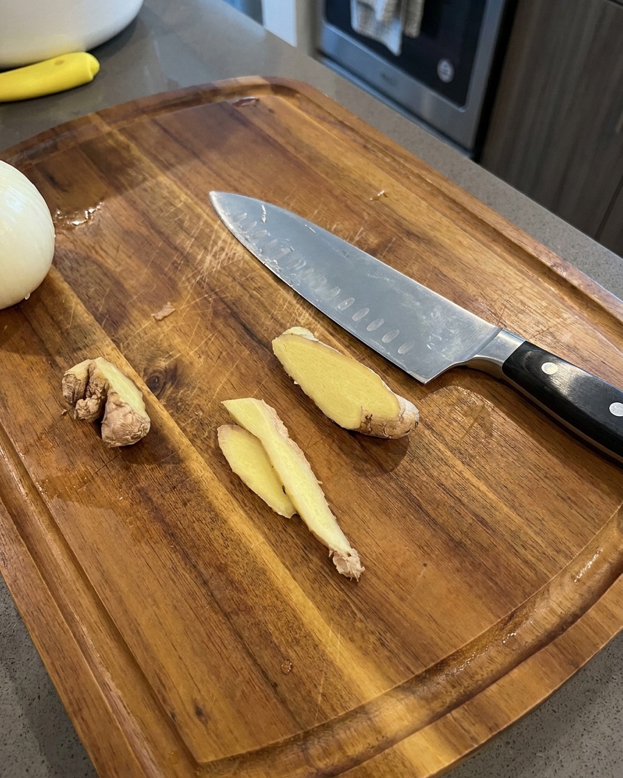 Thin slices of fresh ginger resting on a wooden cutting board next to a chefs knife and an onion half.