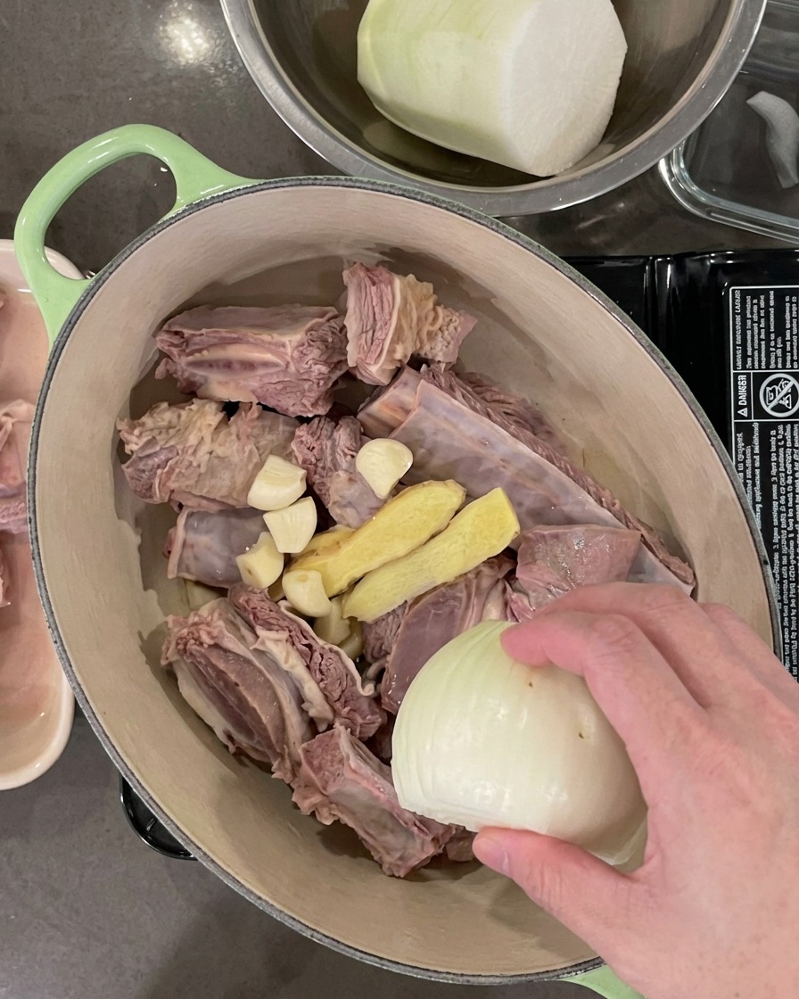 The pot containing the short ribs now includes garlic cloves, ginger slices, an onion half, and green onions.
