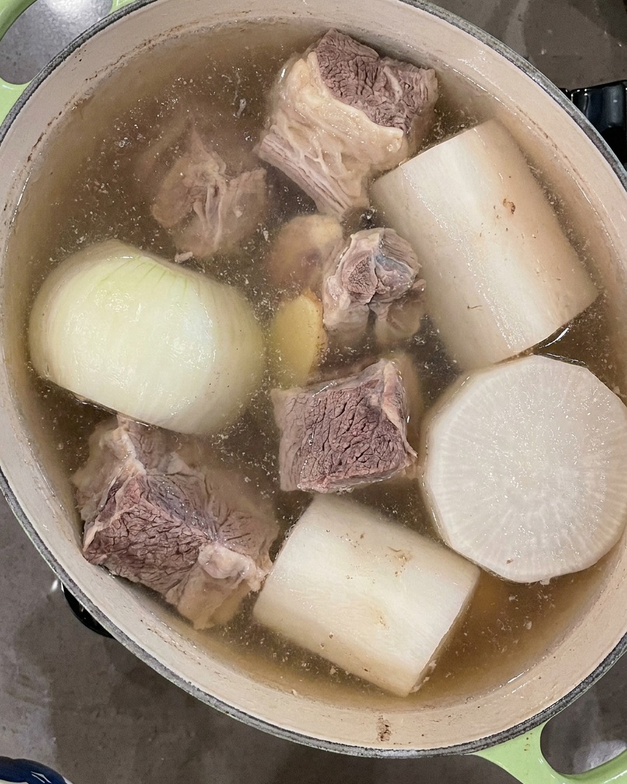 A black ladle skimming floating foam off the surface of the bubbling soup broth.