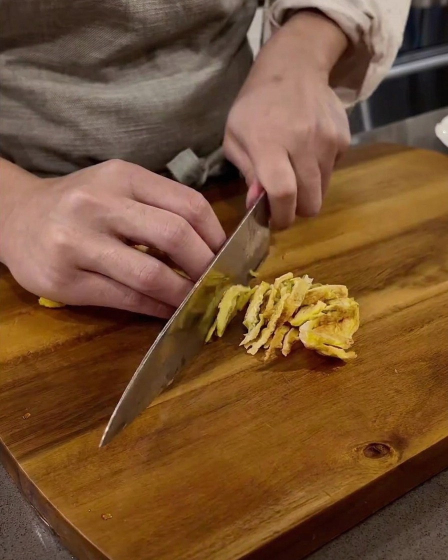 Thin strips of cooked egg being sliced on a wooden cutting board.