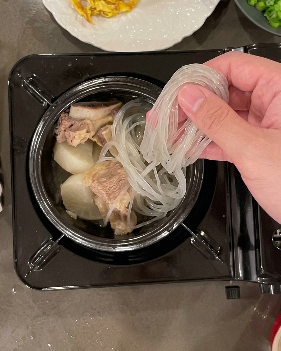 A hand placing a bundle of soaked, translucent glass noodles into a stone pot filled with beef and radish pieces.