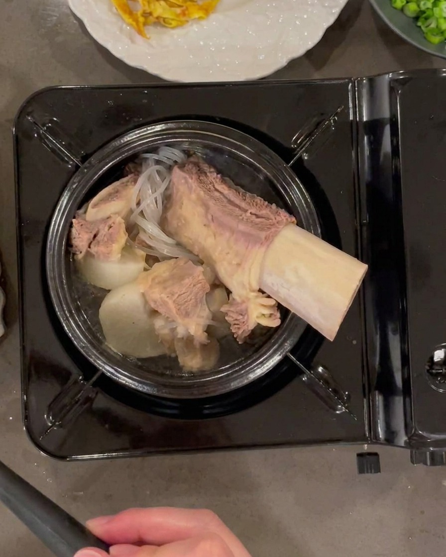 A ladle pouring hot, clear beef broth into a stone pot filled with beef, radish, and glass noodles.