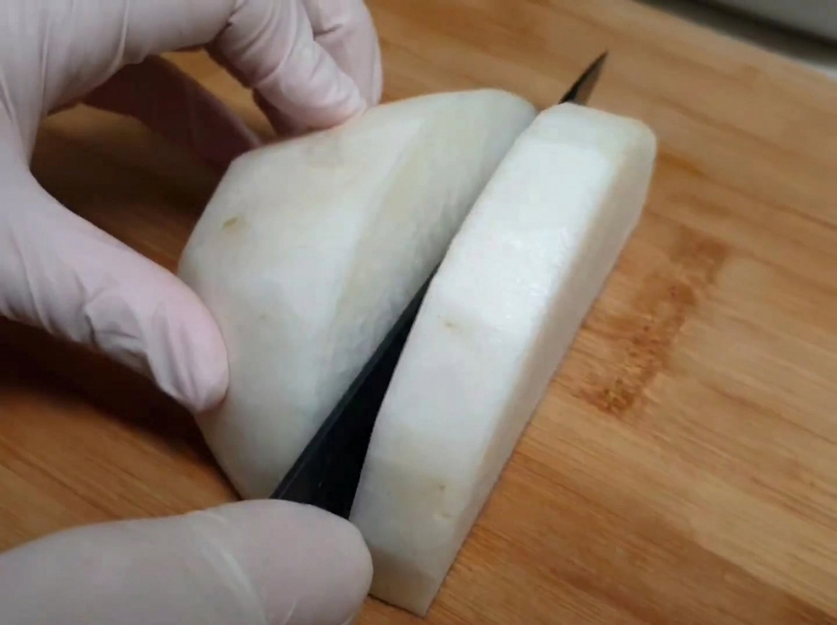 White radish being sliced on a wooden board with a kitchen knife, hands wearing food-safe gloves.
