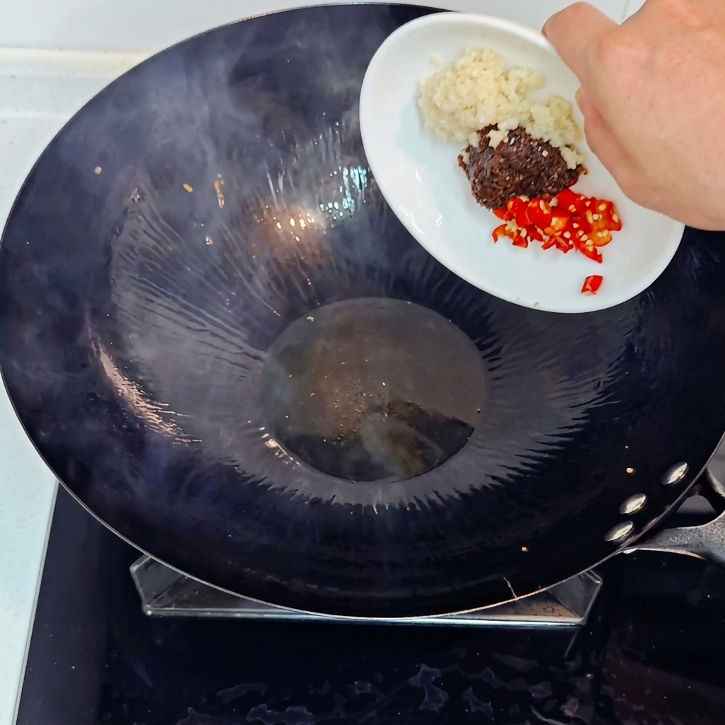 Adding minced garlic, chopped red chili, and soybean paste into a hot wok from a small white plate.