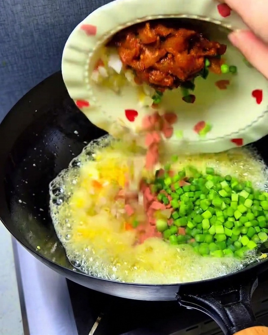 Chopped kimchi, diced ham, chicken, and green garlic sprouts being poured from a plate into the wok with the eggs.