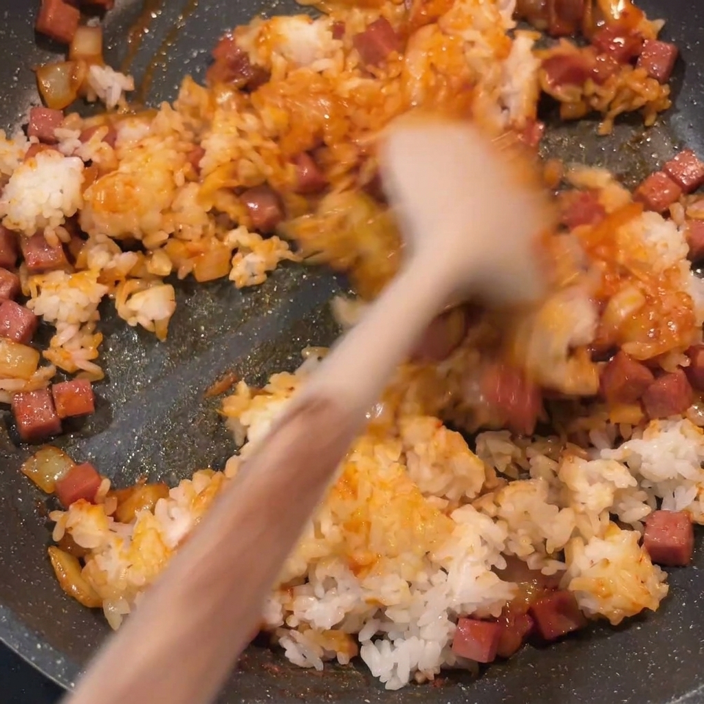 The finished kimchi fried rice being stirred in a pan, showing a consistent red color and well-distributed pieces of spam and onion.