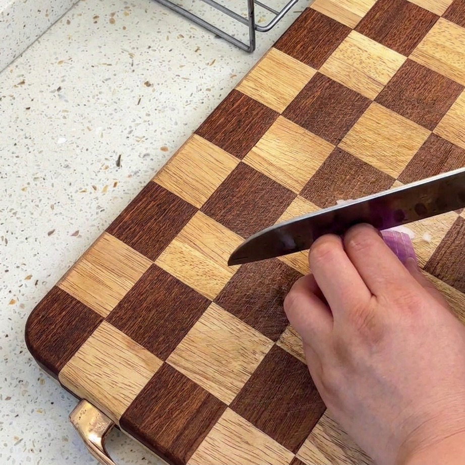 A close-up view of shallots being finely chopped on a checkered wooden cutting board with a sharp knife.
