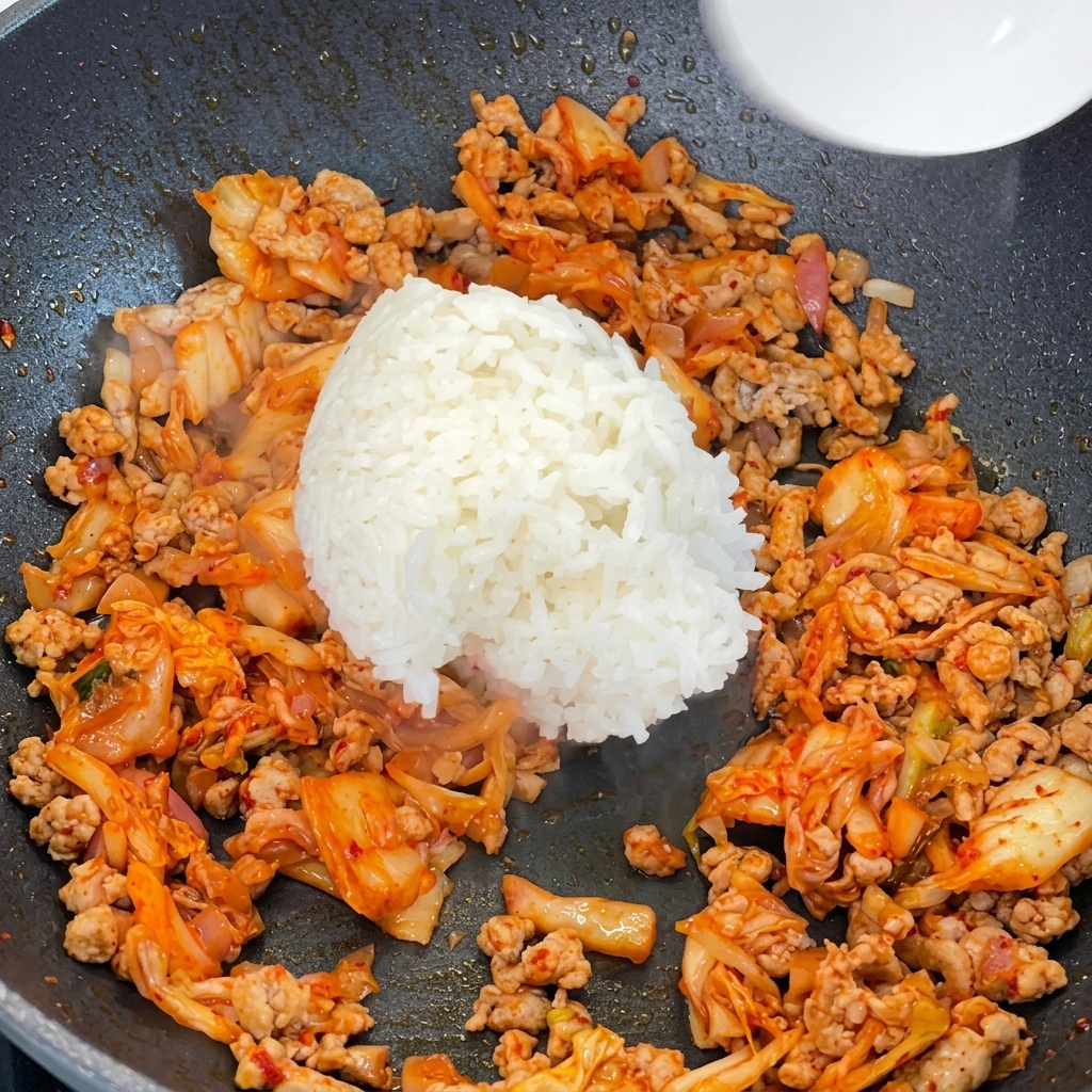 Cooked white rice being added to a pan filled with stir-fried minced pork and kimchi.