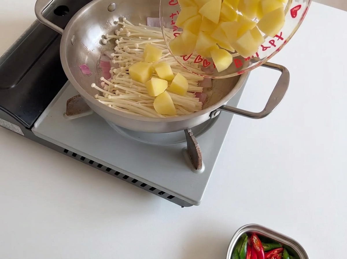 Chunks of raw yellow potato being added to the pan, resting on top of the white enoki mushrooms.