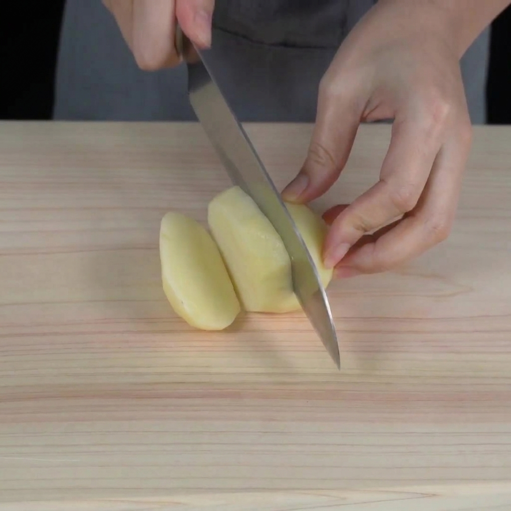 Hands using a chefs knife to cut a peeled raw potato into small cubes on a wooden cutting board.
