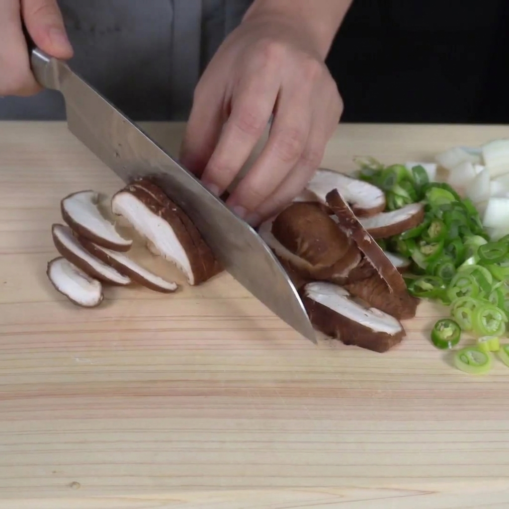 Thinly slicing fresh shiitake mushrooms on a cutting board, with chopped vegetables visible nearby.