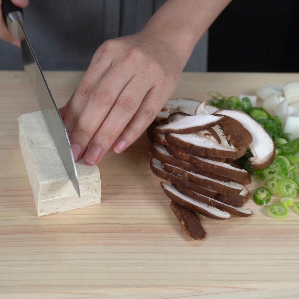 Hands slicing a block of firm white tofu into even, bite-sized cubes on a cutting board.