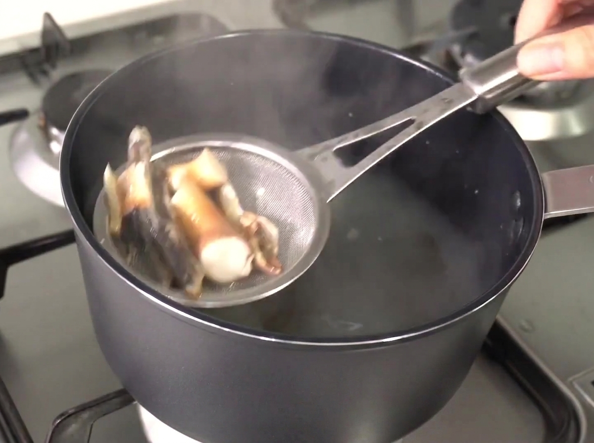 A metal strainer lifting softened anchovies and kelp out of the hot, clear broth.