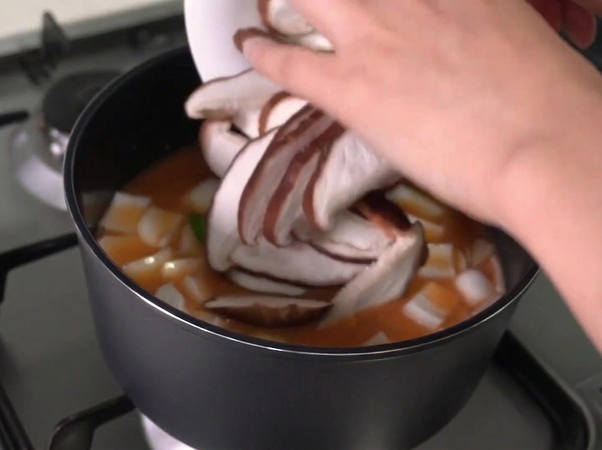 Sliced shiitake mushrooms being added from a white plate into a pot of boiling stew.