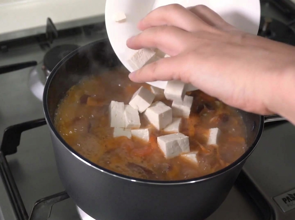 Fresh tofu cubes being added from a white plate into an actively boiling stew.