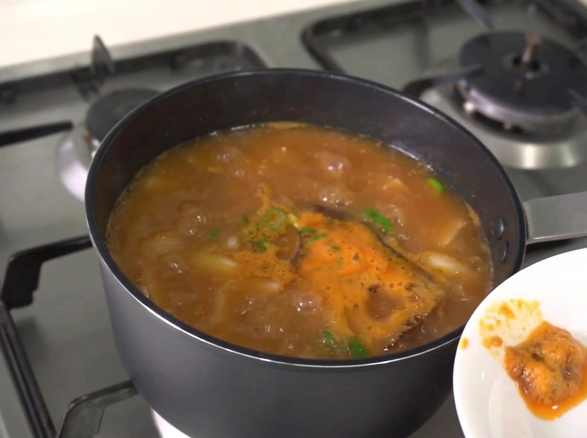 A wooden spoon skimming off the reddish-brown foamy scum from the surface of an actively boiling stew.