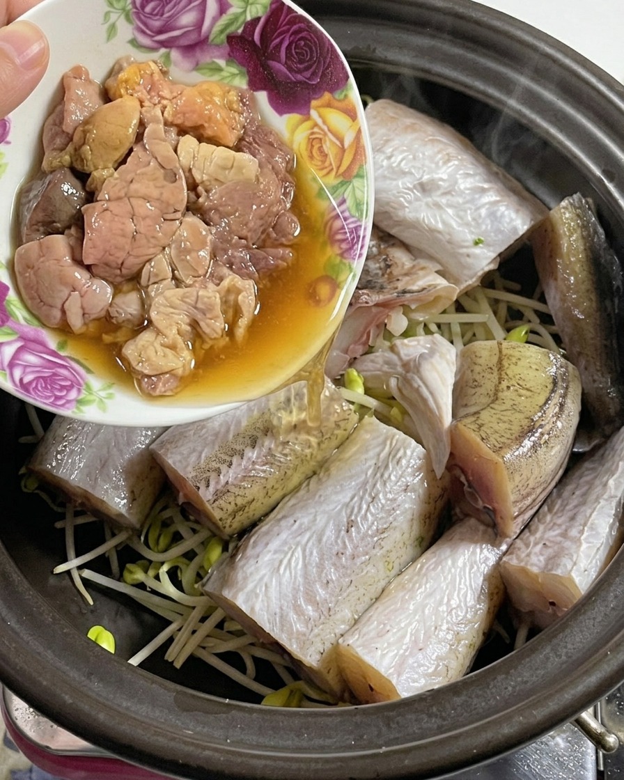 A small floral bowl pouring fresh, delicate pollock roe into a clay pot already containing fish and bean sprouts.