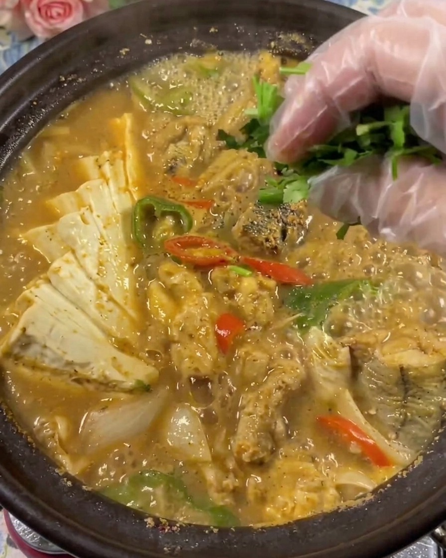 A close-up of a hand wearing a clear plastic glove placing fresh green cilantro into a bubbling clay pot of pollock roe and tofu soup.cite: 1