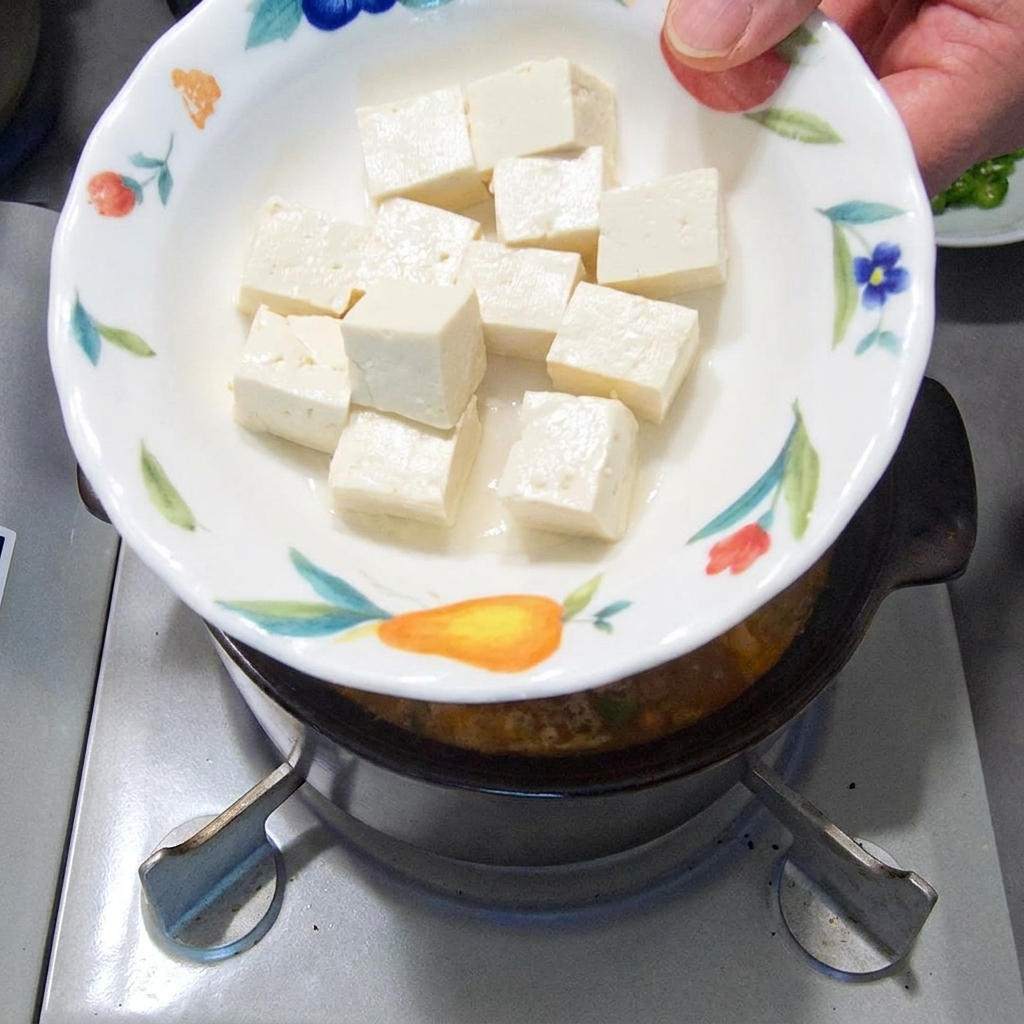 Soft, white tofu cubes being dropped into the simmering stew.