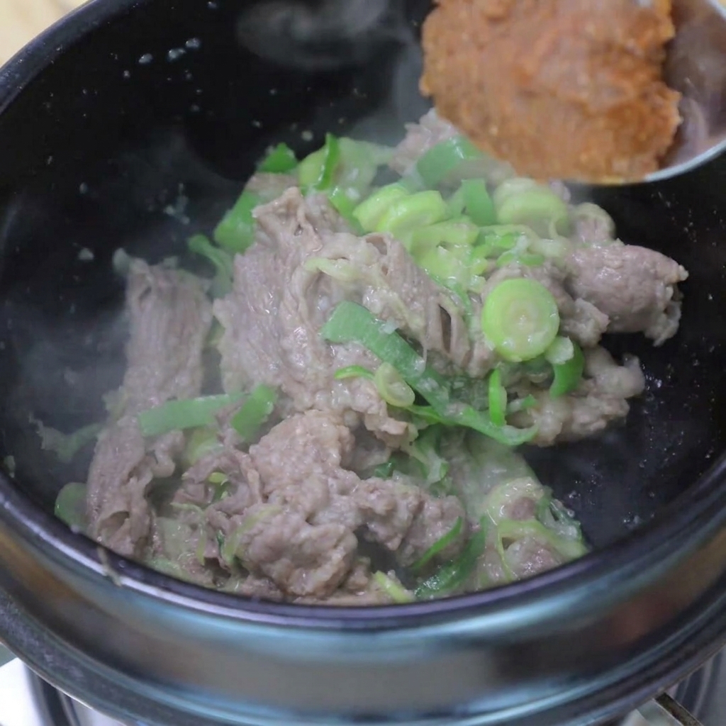 A spoonful of soybean paste being added to the pot containing cooked beef and sautéed green onions.