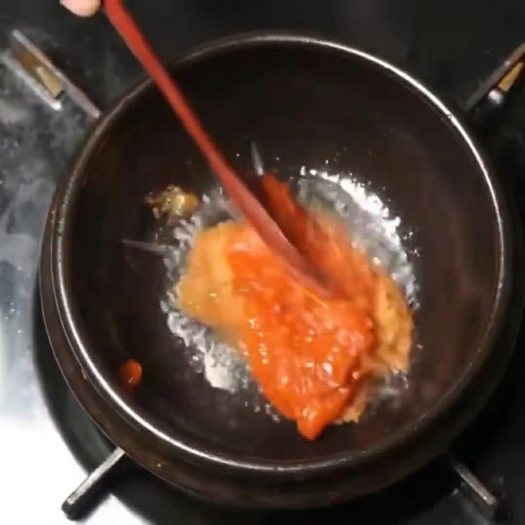 Thick red and brown paste mixture being stirred with a wooden spoon in a black pot over low heat.