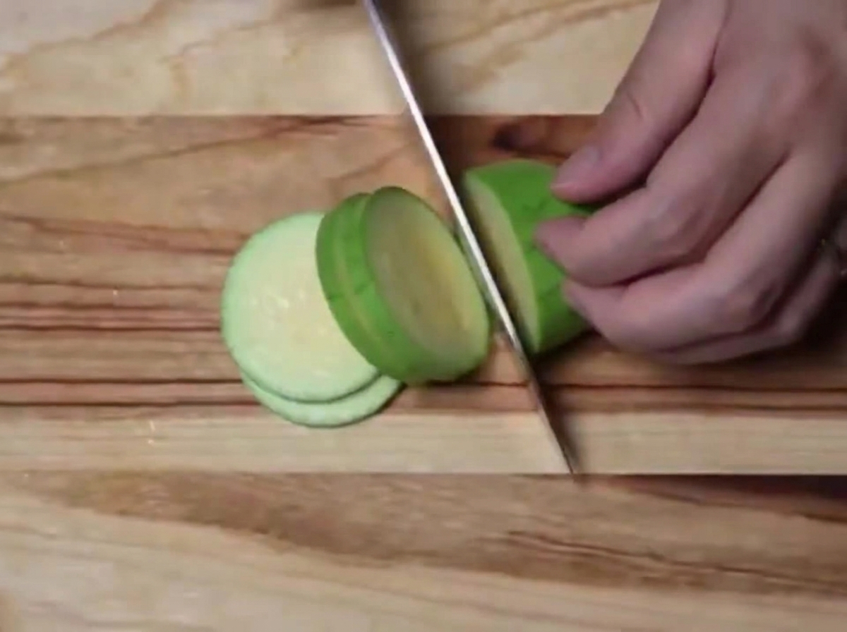 A green zucchini being sliced into uniform semi-circles on a wooden cutting board.