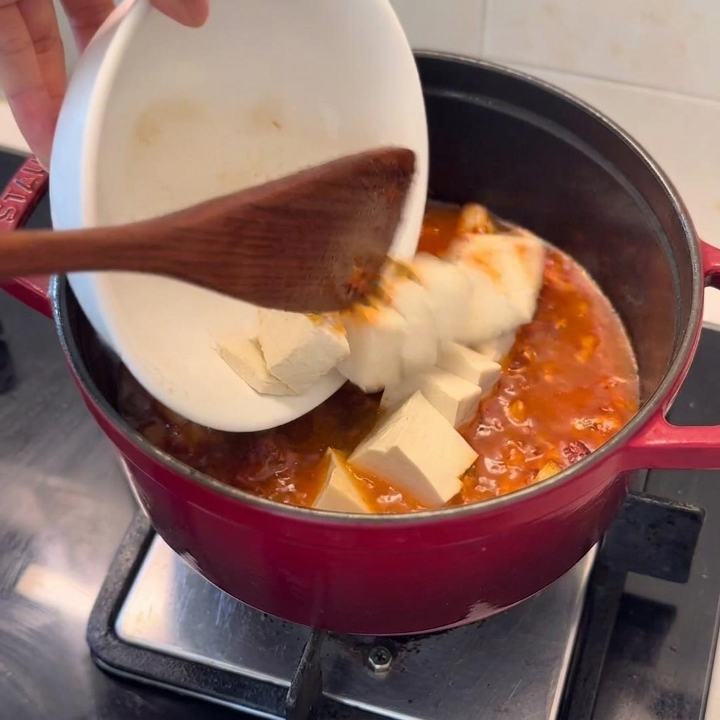 Fresh tofu cubes being carefully poured from a white bowl into a deep red pot of bubbling kimchi pork stew.