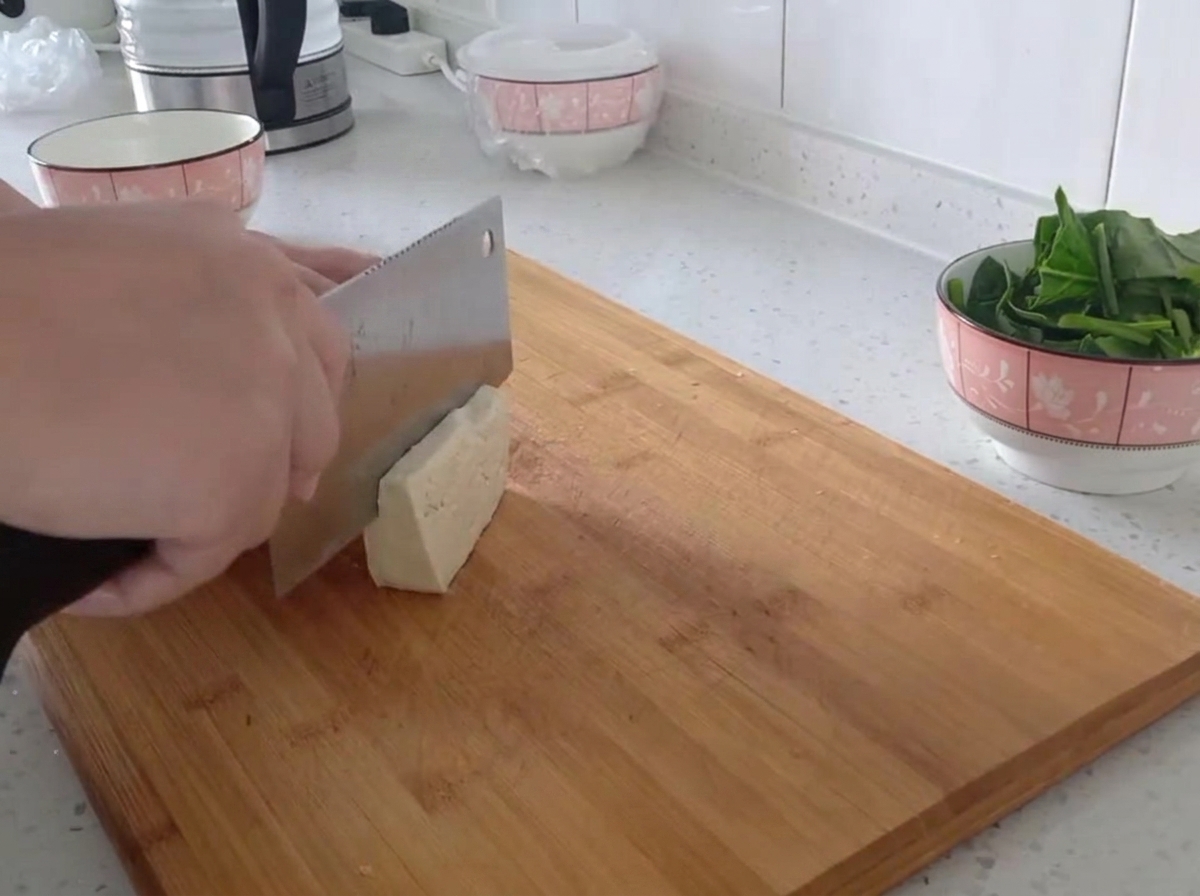 Firm tofu block being cut into thin, even strips on a wooden cutting board.