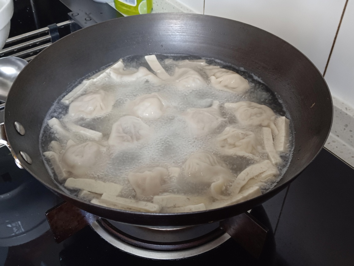 A close-up of vermicelli noodles being submerged into the boiling pot with dumplings and tofu strips.