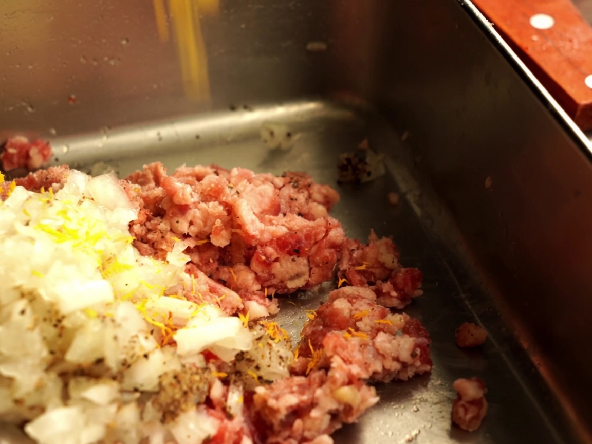 Minced onions and breadcrumbs being added to raw ground beef in a metal container.