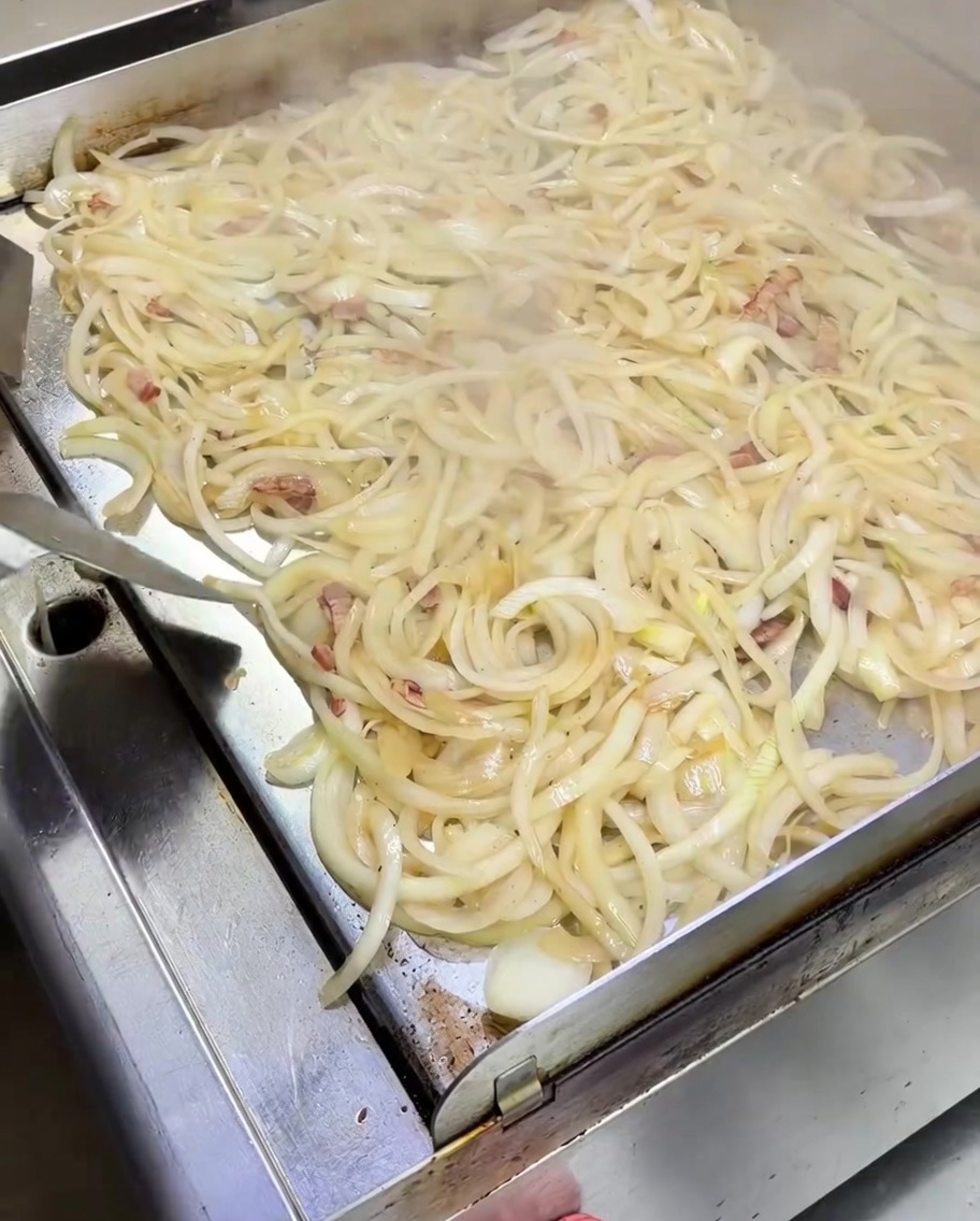Spatula stirring a large mound of softening white onions mixed with bacon bits on a steaming metal griddle.