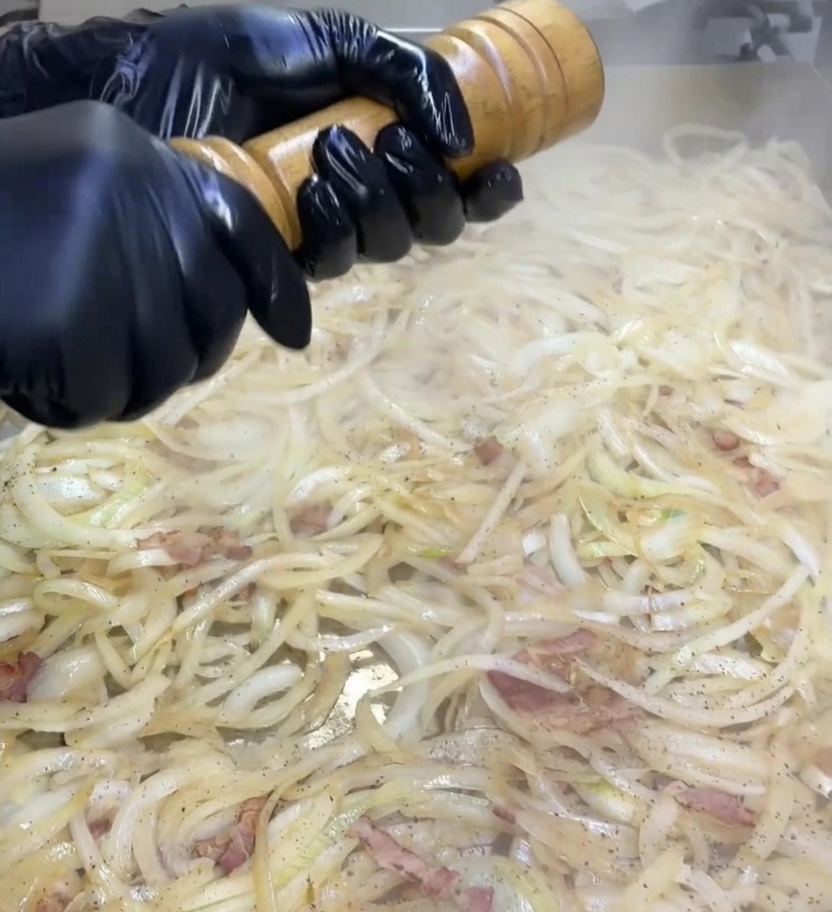 Hands in black gloves twisting a wooden pepper grinder over a griddle covered in cooking onions and bacon.