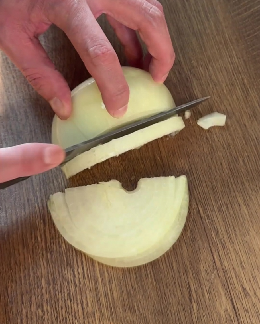 A hand using a knife to slice an onion into thin strips on a wooden cutting board.