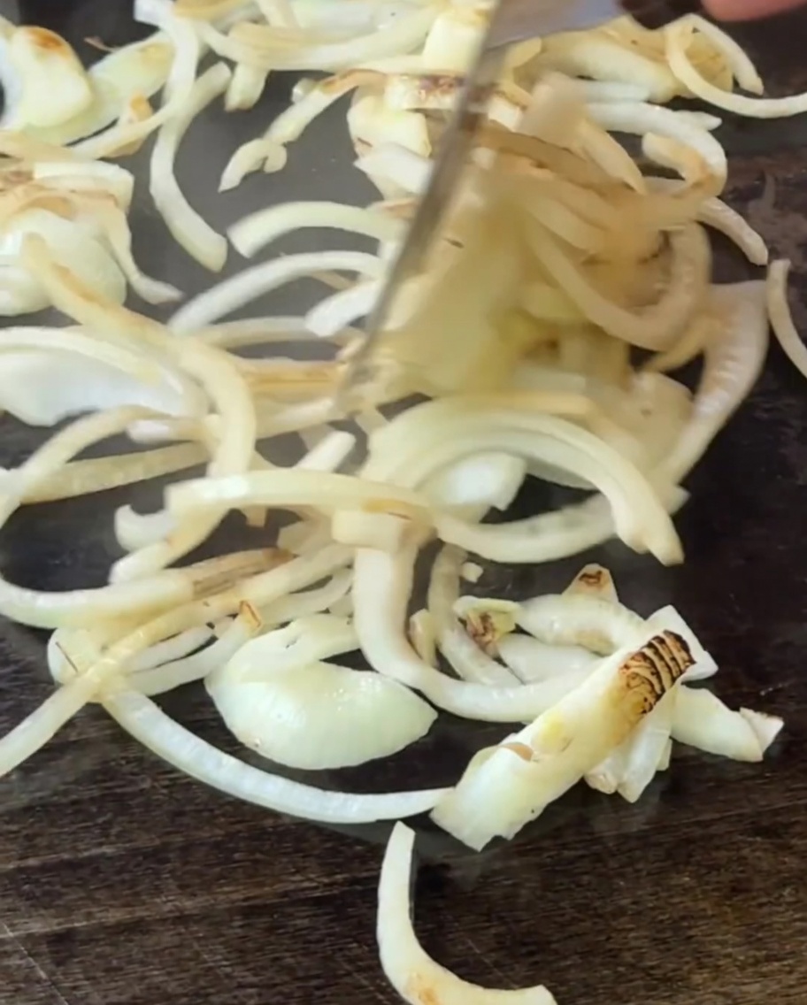 Onions being stirred on the griddle with a spatula as they begin to soften and cook.