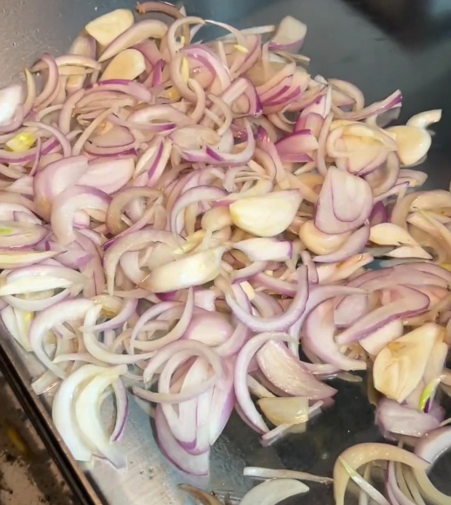 Sliced onions being stirred on a hot griddle with a metal spatula.