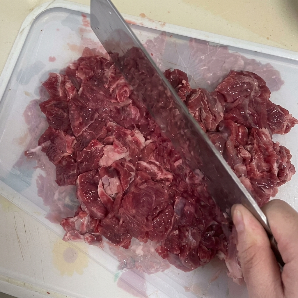 A cleaver being used to mince fresh beef on a cutting board.