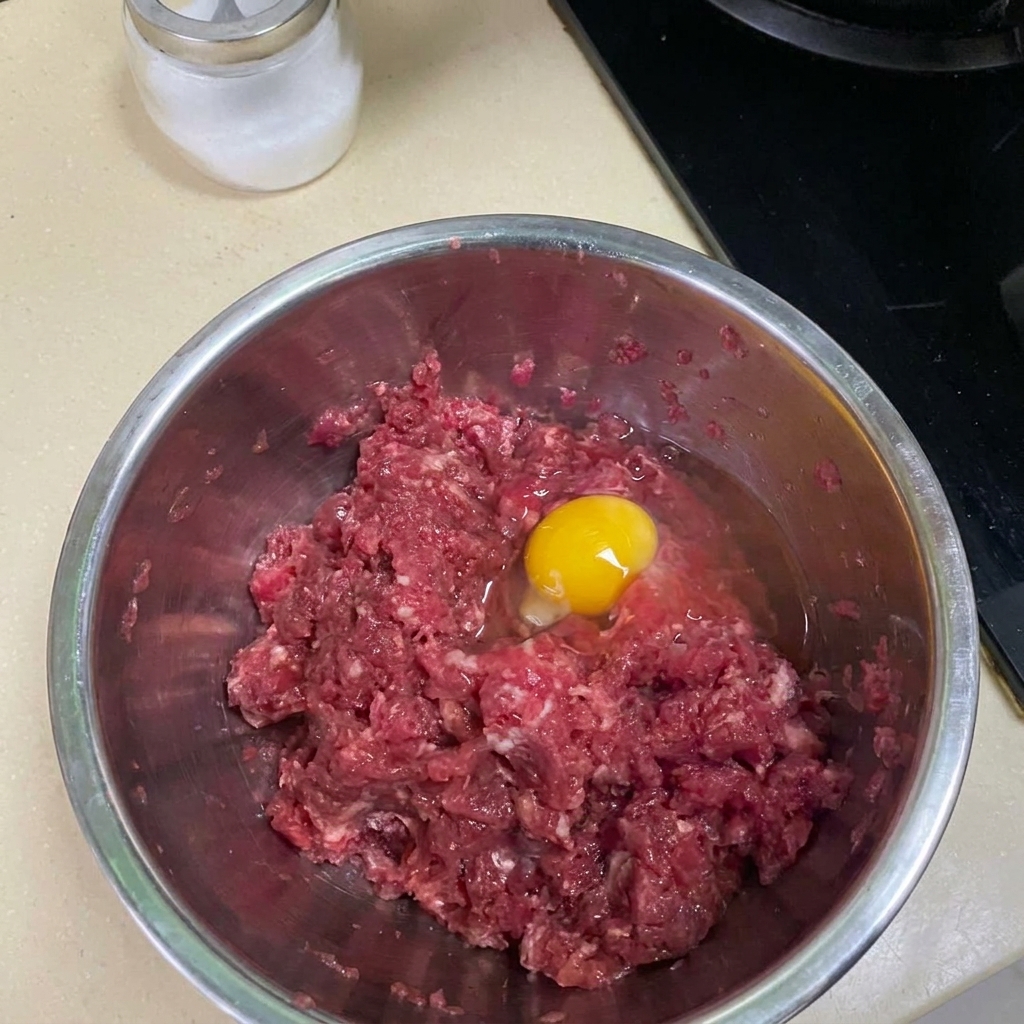 Ground beef being mixed with spices and an egg in a metal bowl.