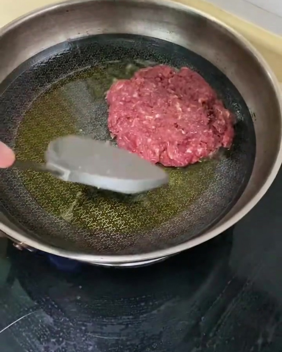 A seasoned beef patty frying in a hot skillet with oil.